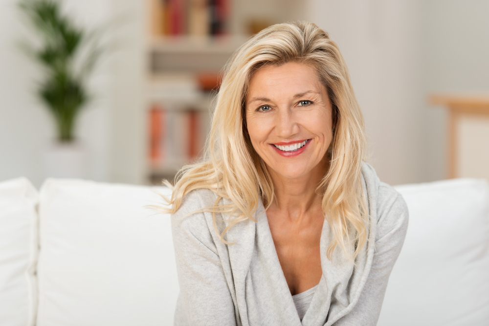 Lovely middle-aged blond woman with a beaming smile sitting on a sofa at home looking at the camera Lovely middle-aged blond woman with a beaming smile sitting on a sofa at home looking at the camera